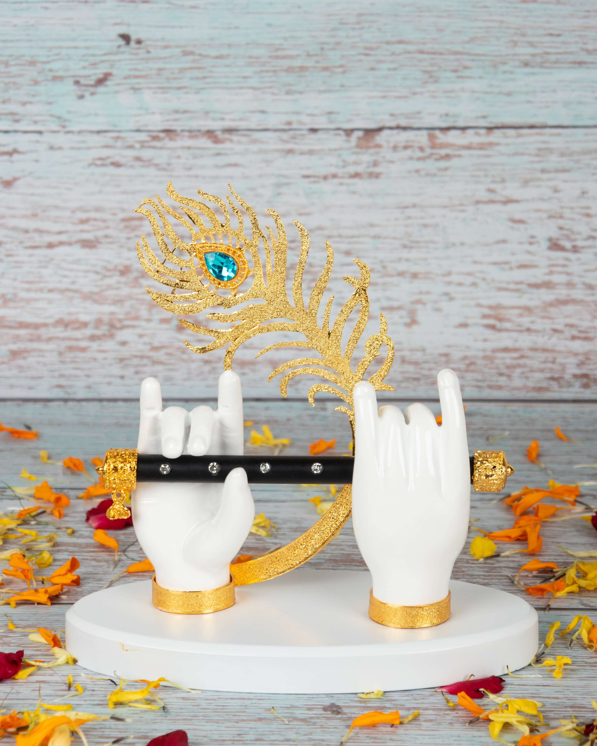An idol depicting Lord Krishna's hands in white, holding a black flute with golden accents, and a prominent golden peacock feather with a blue gem at its eye, all resting on a white base amidst scattered orange and red flower petals, set against a light blue wooden background.