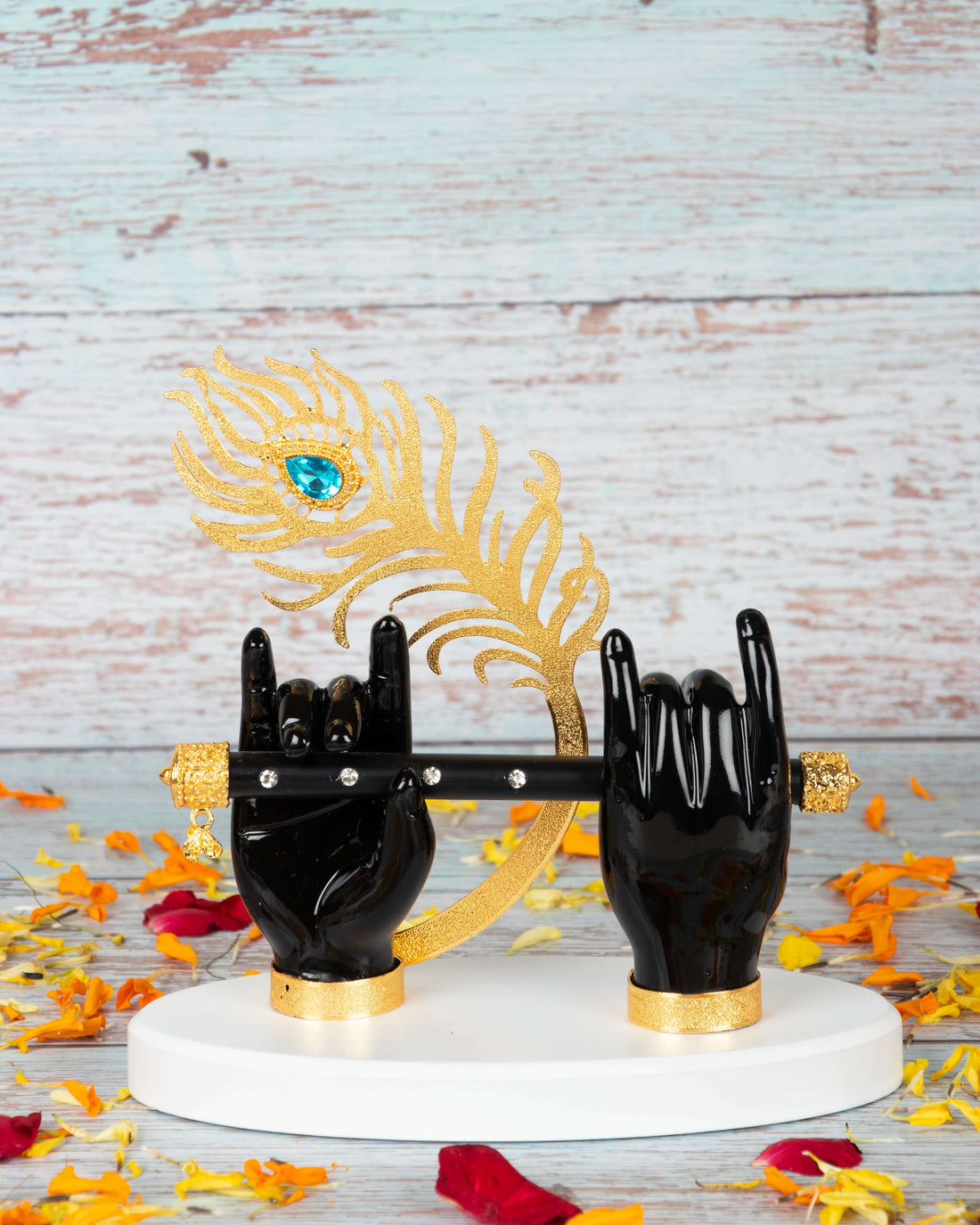 A decorative idol featuring two black hands of Lord Krishna holding a black flute, adorned with a golden peacock feather with a blue jewel and resting on a white and gold base. The idol is held by a person with manicured hands, against a blurred background of light-colored wooden planks.