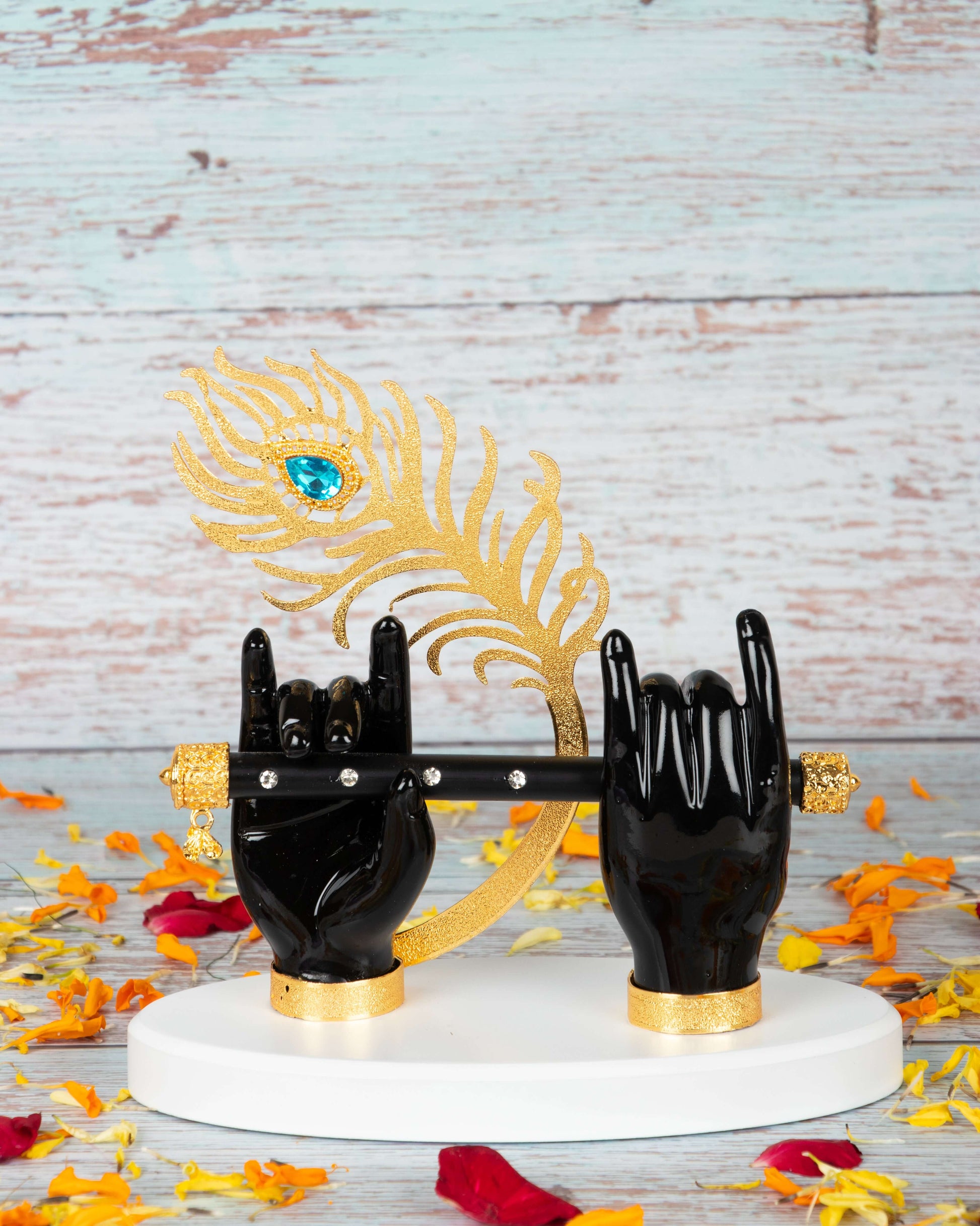 A decorative figurine depicting Lord Krishna's black hands adorned with golden accents, holding a black flute, with a golden peacock feather featuring a blue gem, all resting on a white base and surrounded by scattered orange and red flower petals, set against a light wooden background.