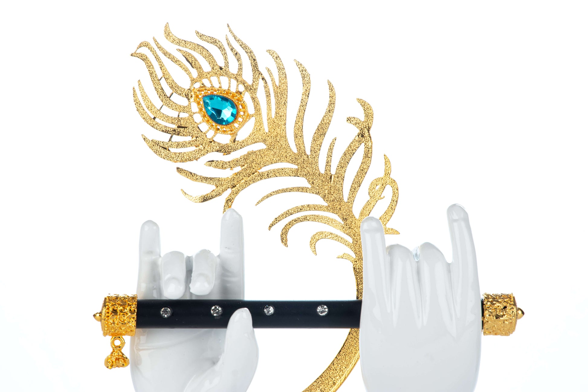 A decorative figurine depicting two white sculpted hands, representing Lord Krishna's hands, holding a black and gold flute adorned with jewel-like accents. Behind the hands and flute, a large, ornate golden peacock feather with a prominent blue-green gemstone in its eye is positioned on a white background.