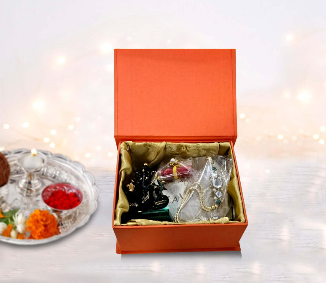 An open red gift box displaying a beautifully designed envelope, a traditional Rakhi, Roli Chawal, and a crystal Ganesha idol. Handmade chocolates are also visible.