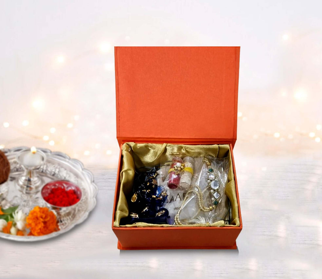 An open red gift box displaying a beautifully designed envelope, a traditional Rakhi, Roli Chawal, and a crystal Ganesha idol. Handmade chocolates are also visible.