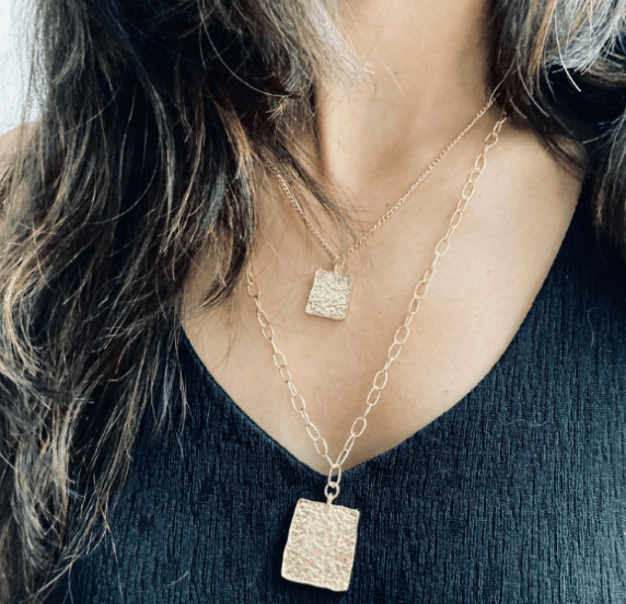 A person is wearing a layered necklace set. The set includes a shorter, finer chain with a small square metal pendant and a longer, larger chain with a larger square metal pendant. Both pendants have a textured or hammered finish.