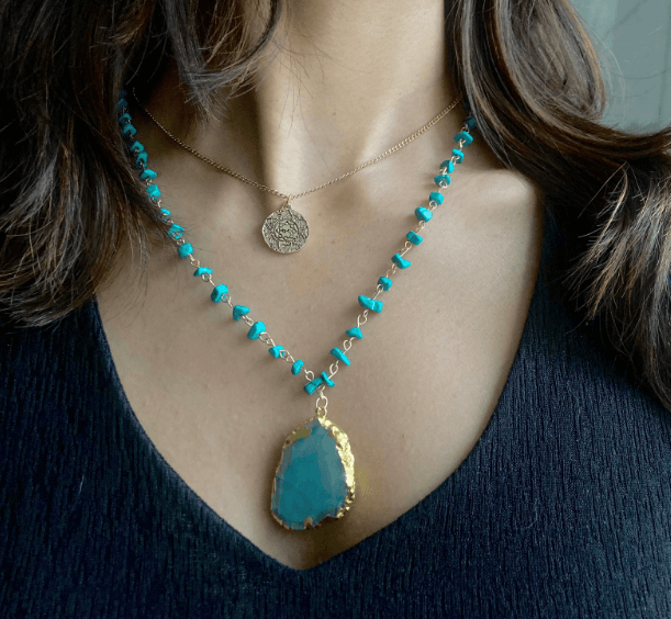 A layered Agate stone pendant necklace is worn by a woman over a dark top. The longer necklace features small, irregular-shaped turquoise-colored beads connected by a gold-toned chain, with a large, similarly colored agate stone pendant with a gold-plated edge. The shorter necklace is a simple gold-toned chain with a small, circular coin-like pendant.