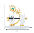 A Lord Krishna hand idol featuring two white hands adorned with gold-plated bracelets, holding a black flute, with a vibrant gold and blue peacock feather behind it. The idol stands on a white base, and the image includes annotations indicating its dimensions: 7 inches in height, 7 inches in width, and 4 inches in depth.