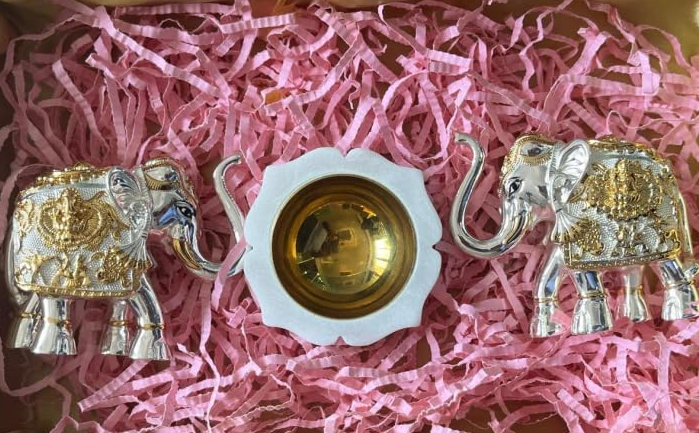 Two silver-plated, trunk-up elephant statues with a marble diya and brass accents, presented on a pink bed of rose petals.