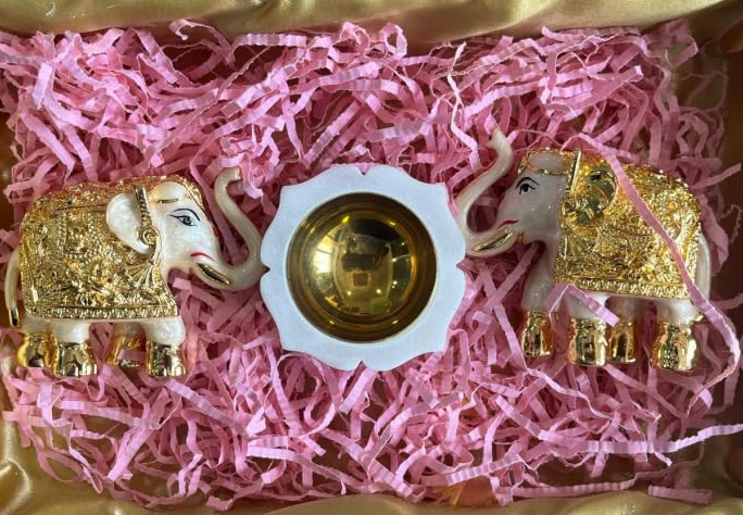 A set of two golden trunk-up elephant idols on a bed of pink tissue paper, with a marble diya with brass accents in the center.