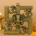 Brass-finished decorative swastika wall hanging with embossed Om symbols and a central Lord Ganesha figure, placed against a warm background with a brass oil lamp and marigold flowers.