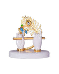A decorative idol featuring Lord Krishna's white hands holding a golden flute, with a golden peacock feather and a miniature blue figure of Krishna playing the flute on a white base.