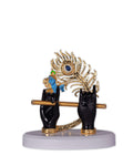 A decorative idol featuring two black hands holding a golden flute with a large golden peacock feather and a small blue figure of Lord Krishna, all resting on a white base.