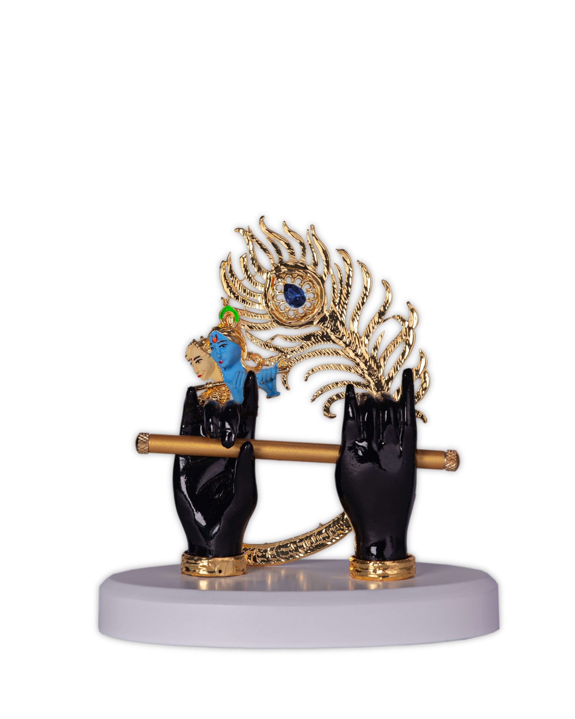 A decorative idol featuring two black hands holding a golden flute with a large golden peacock feather and a small blue figure of Lord Krishna, all resting on a white base.
