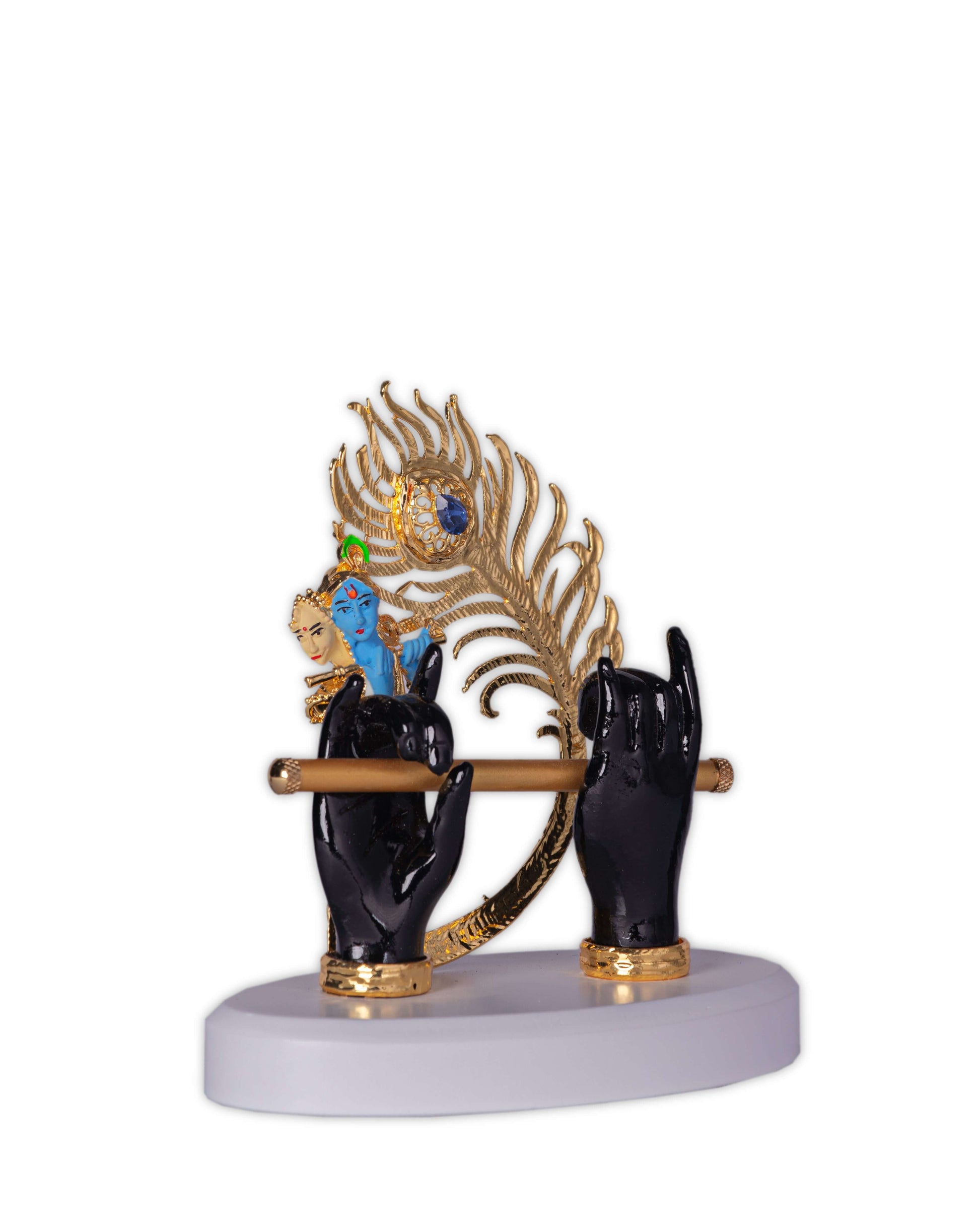 A decorative idol depicting two black hands holding a golden flute, adorned with a large golden peacock feather and a small blue figure of Lord Krishna, all set on a white base.