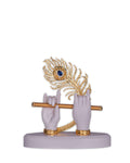 A decorative white idol featuring Lord Krishna's hands holding a golden flute, with a golden peacock feather adorned with a blue gem resting between the hands, all set on a light-colored base.