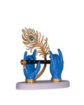 A decorative figurine featuring two blue hands, stylized as Lord Krishna's, holding a black flute horizontally. Behind the flute, a detailed golden peacock feather adorned with a blue jewel is positioned. The entire sculpture rests on a light-colored, possibly white or grey, base.
