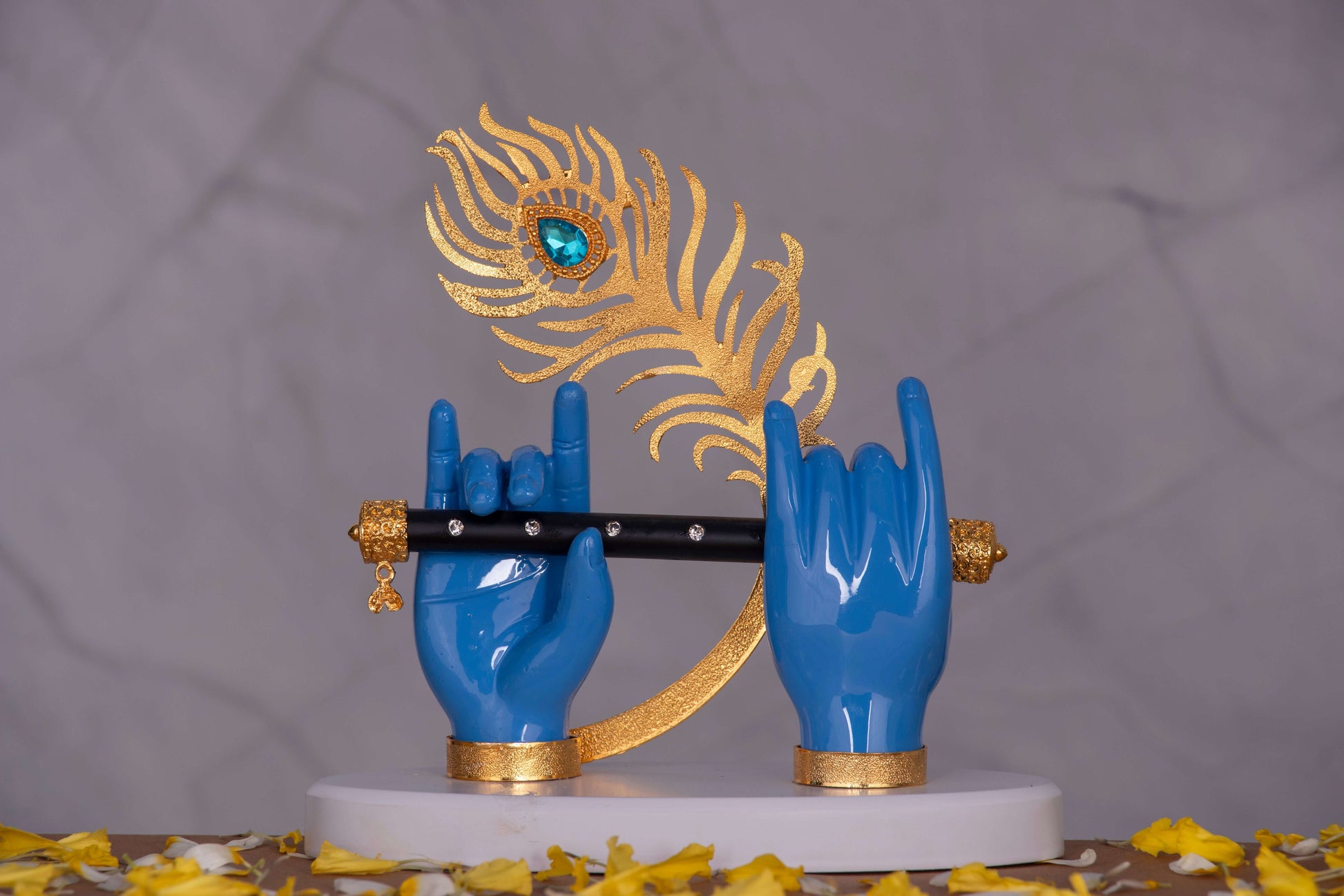A close-up shot of a stylized Lord Krishna hands statue, featuring blue hands holding a black flute with golden ends, positioned against a large golden peacock feather adorned with a blue jewel, all resting on a white base with scattered yellow petals around it.
