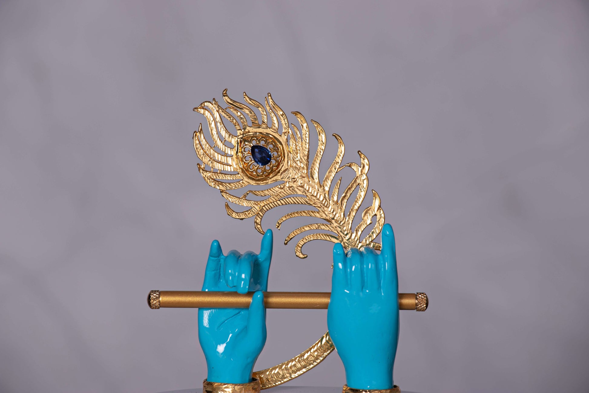 A striking sculpture featuring two blue hands, emblematic of Lord Krishna, holding a golden flute, with a majestic golden peacock feather adorned with a prominent blue gem positioned behind them.