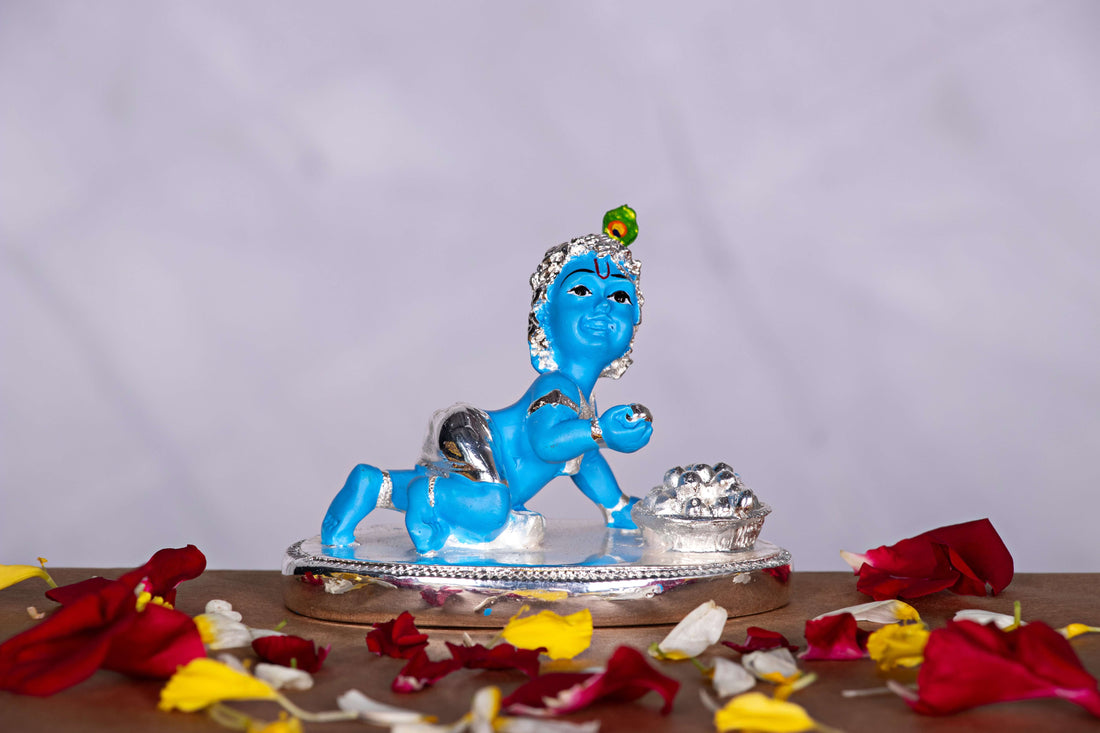 A statue of Bala Krishna (Baby Krishna) crawling, depicted with blue skin, silver-colored clothing, and a peacock feather on his head, with a small pot of butter next to him.