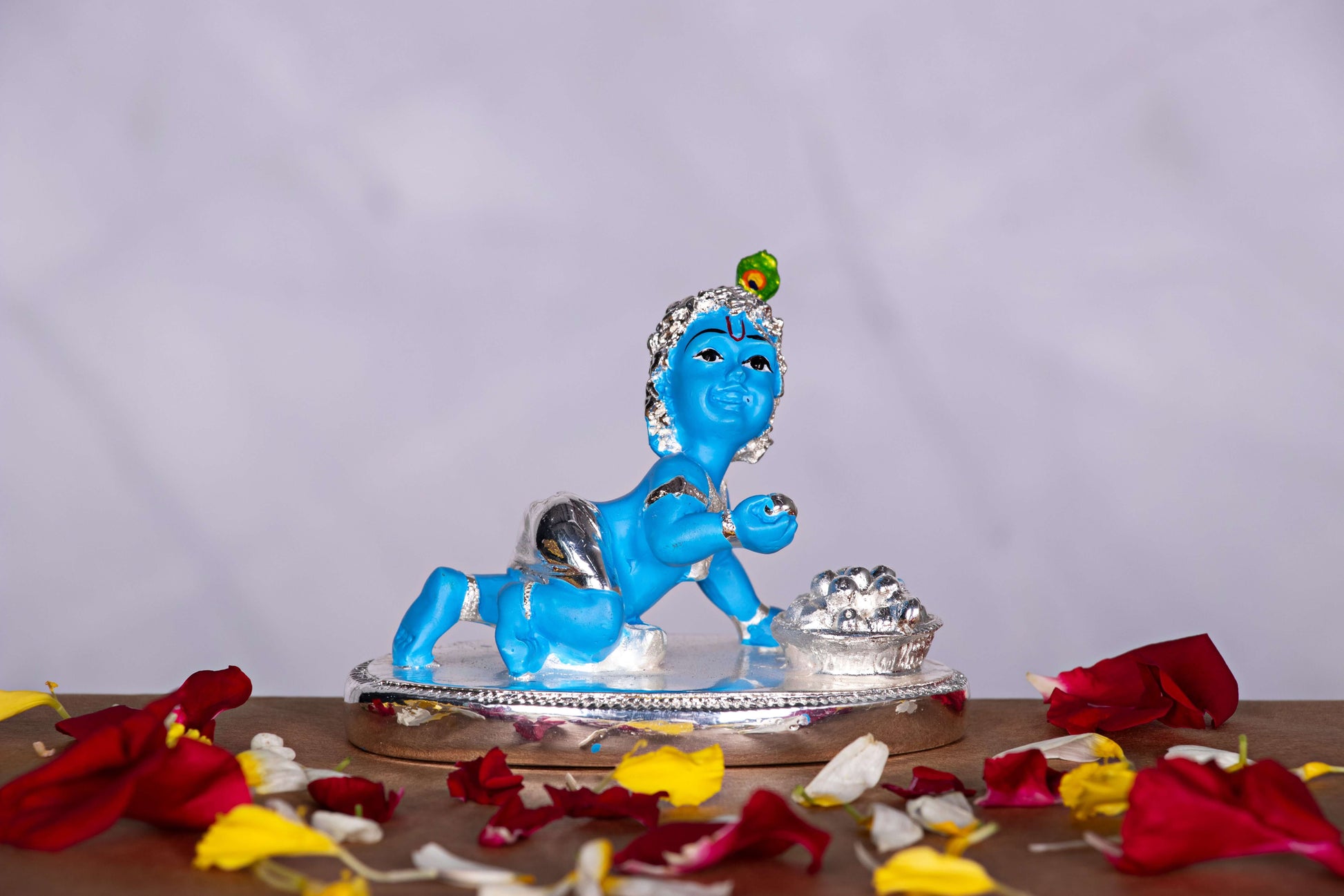 Blue and silver baby Krishna idol in a crawling pose with butter pot, decorated with peacock feather on head, surrounded by scattered flower petals.