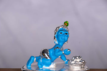A statue of Bala Krishna (Baby Krishna) crawling, depicted with blue skin, silver-colored clothing, and a peacock feather on his head, with a small pot of butter next to him.