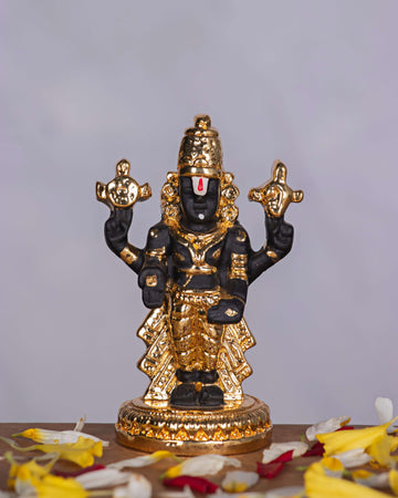 meticulously crafted black and gold-toned idol of Lord Venkateswara, also known as Tirupati Balaji, adorned with intricate golden ornaments including a crown, necklaces, armlets, and a base, holding the Shankha (conch) and Chakra (discus) in his upper hands, set against a plain, light-colored background with scattered flower petals at the base.