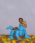 An image of a blue-skinned Bala Krishna (Baby Krishna) idol crawling, adorned with gold jewelry and a peacock feather in his hair, surrounded by yellow flower petals.