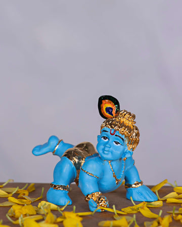 An image of a blue-skinned Bala Krishna (Baby Krishna) idol crawling, adorned with gold jewelry and a peacock feather in his hair, surrounded by yellow flower petals.