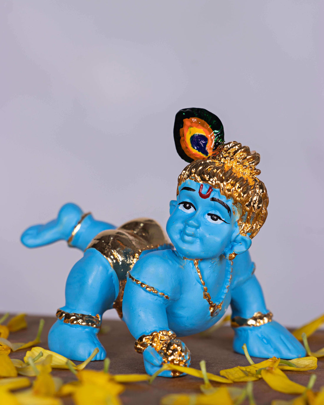 An image of a blue-skinned Bala Krishna (Baby Krishna) idol crawling, adorned with gold jewelry and a peacock feather in his hair, surrounded by yellow flower petals.