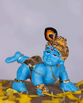 A blue idol of crawling Baby Krishna, also known as Bala Krishna, adorned with golden jewelry and a peacock feather on his head, surrounded by yellow flower petals.