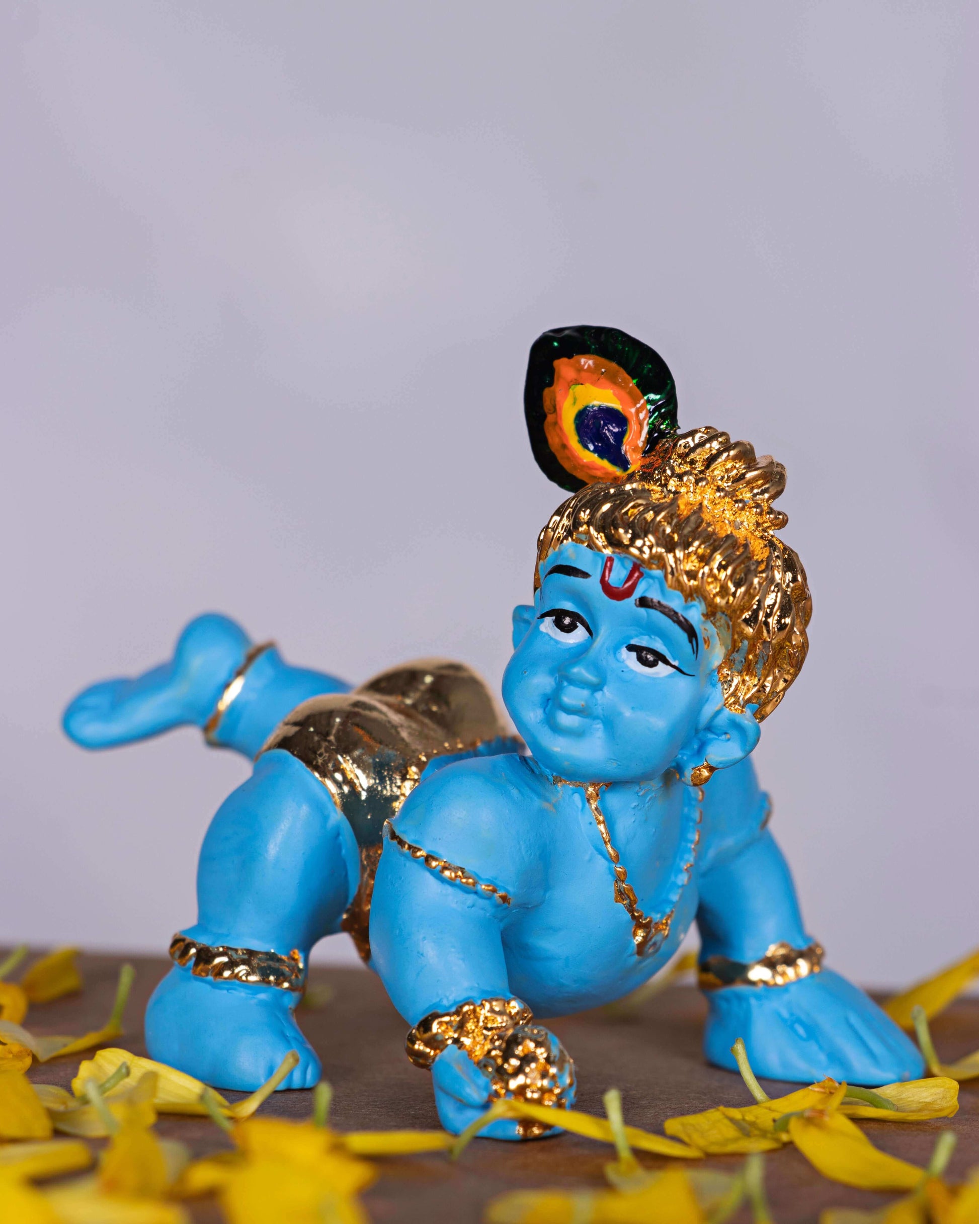 A blue idol of crawling Baby Krishna, also known as Bala Krishna, adorned with golden jewelry and a peacock feather on his head, surrounded by yellow flower petals.
