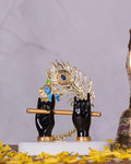 A decorative idol featuring Lord Krishna's black hands holding a golden flute, with a vibrant gold and blue peacock feather rising behind them. The idol stands on a white base, surrounded by scattered yellow flower petals, all set against a softly lit marble-patterned background.