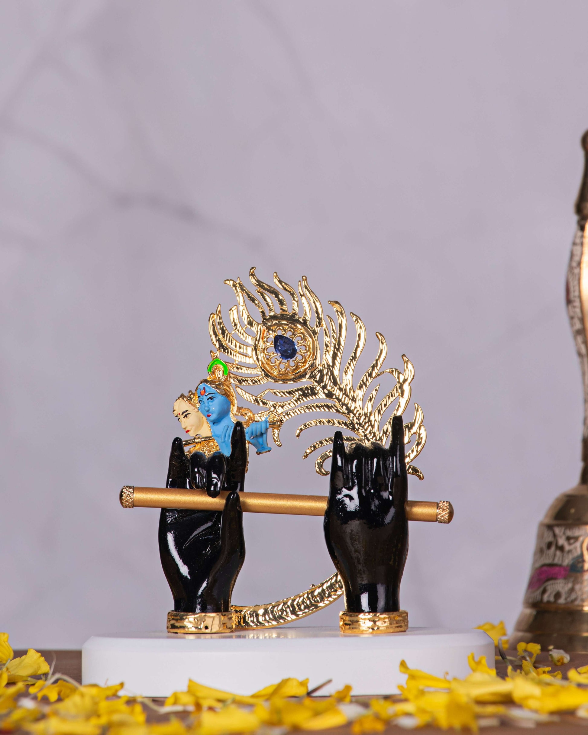 A decorative idol featuring Lord Krishna's black hands holding a golden flute, with a vibrant gold and blue peacock feather rising behind them. The idol stands on a white base, surrounded by scattered yellow flower petals, all set against a softly lit marble-patterned background.