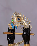 A captivating idol of Lord Krishna, featuring black hands holding a golden flute, set against a large, elaborate golden peacock feather with a blue gem at its center. A small blue-faced Krishna figure is also visible within the feather's design.