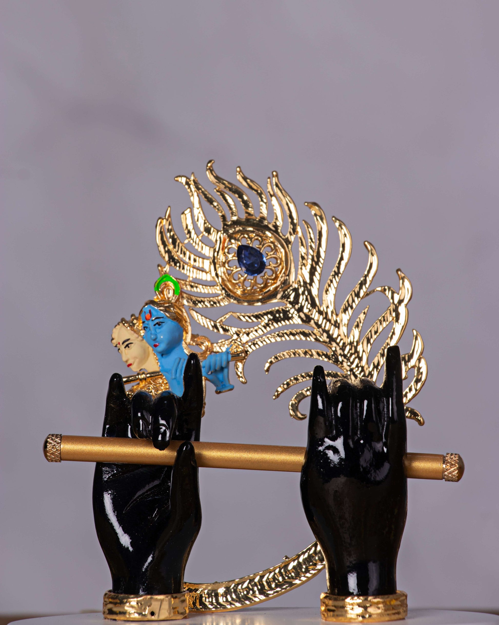 A captivating idol of Lord Krishna, featuring black hands holding a golden flute, set against a large, elaborate golden peacock feather with a blue gem at its center. A small blue-faced Krishna figure is also visible within the feather's design.