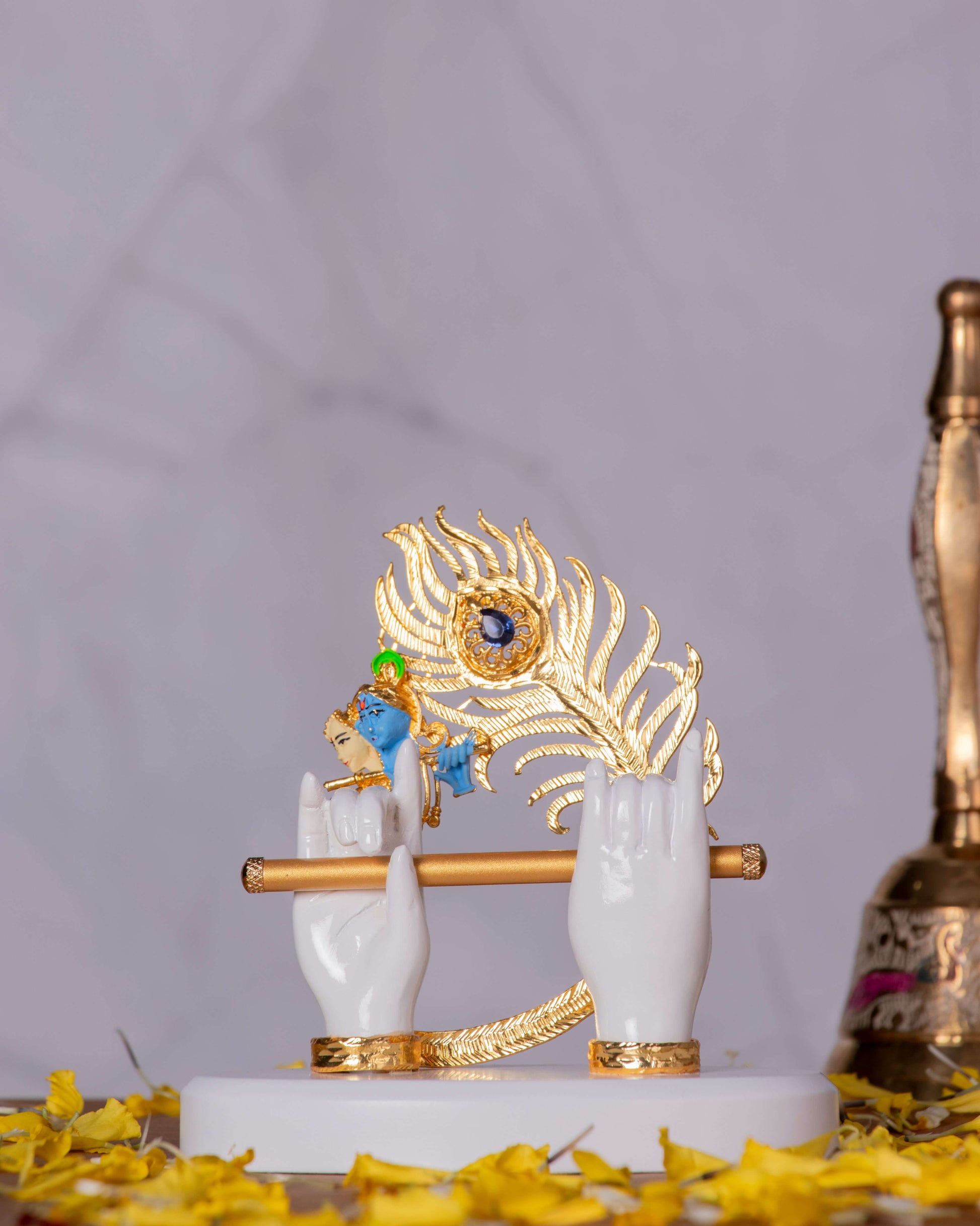 An idol depicting Lord Krishna's divine hands, holding a golden flute, with a small blue face and a golden peacock feather on a white and gold base.