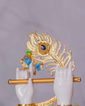 A close-up of a decorative idol featuring two white hands holding a golden flute, with a blue-faced Lord Krishna figure in the background playing it, all set against a large, ornate golden peacock feather with a blue gem at its eye.