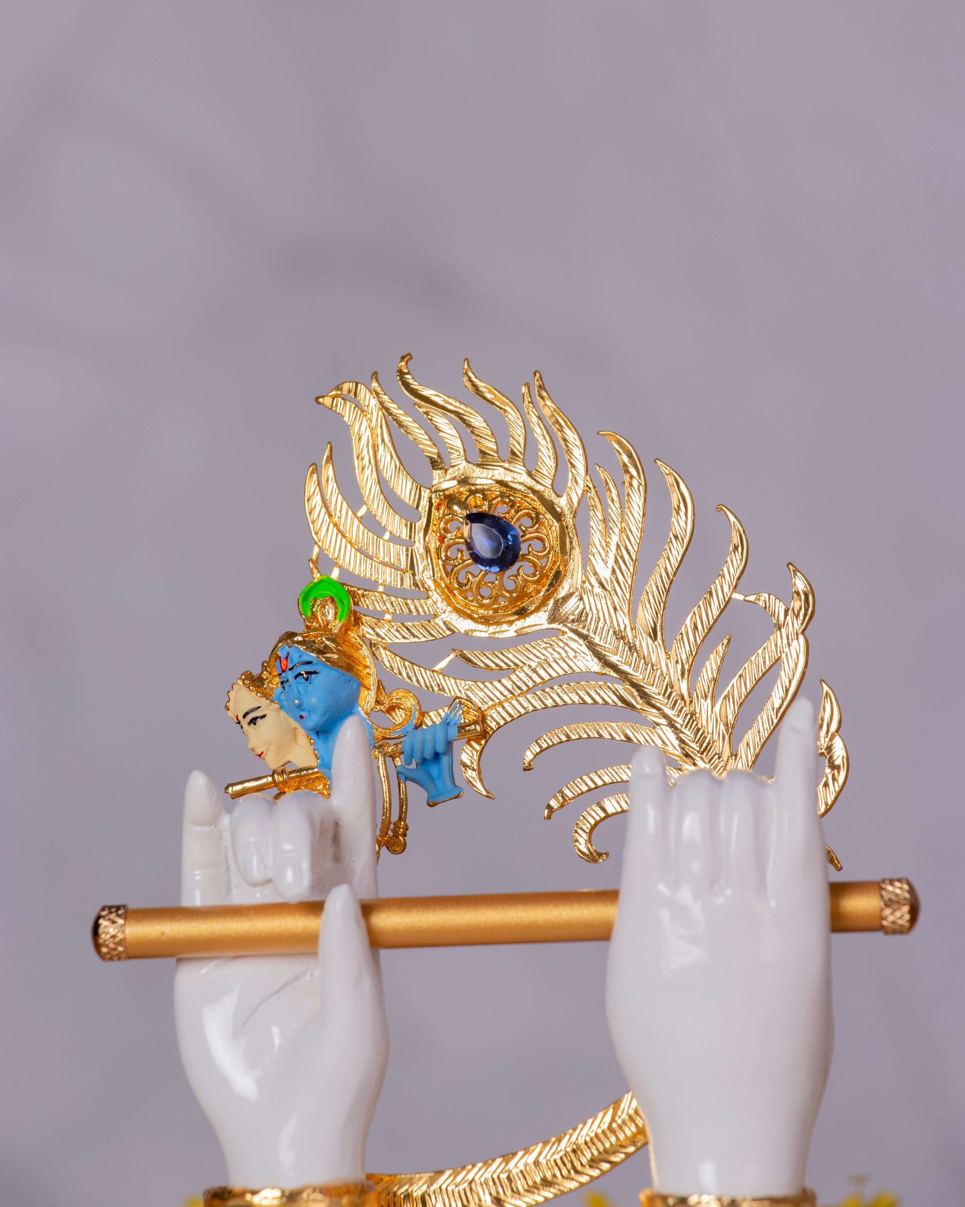 A close-up of a decorative idol featuring two white hands holding a golden flute, with a blue-faced Lord Krishna figure in the background playing it, all set against a large, ornate golden peacock feather with a blue gem at its eye.
