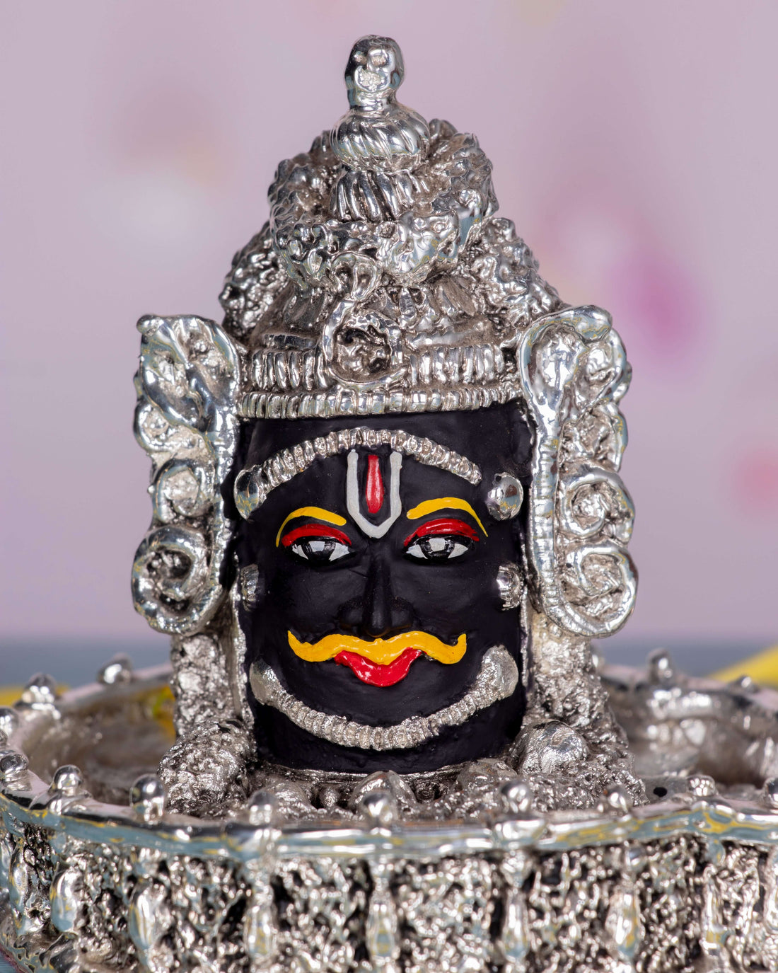 A silver-plated Mahakal idol, a fierce form of Lord Shiva, adorned with intricate details and traditional markings, placed on a surface scattered with yellow flower petals.