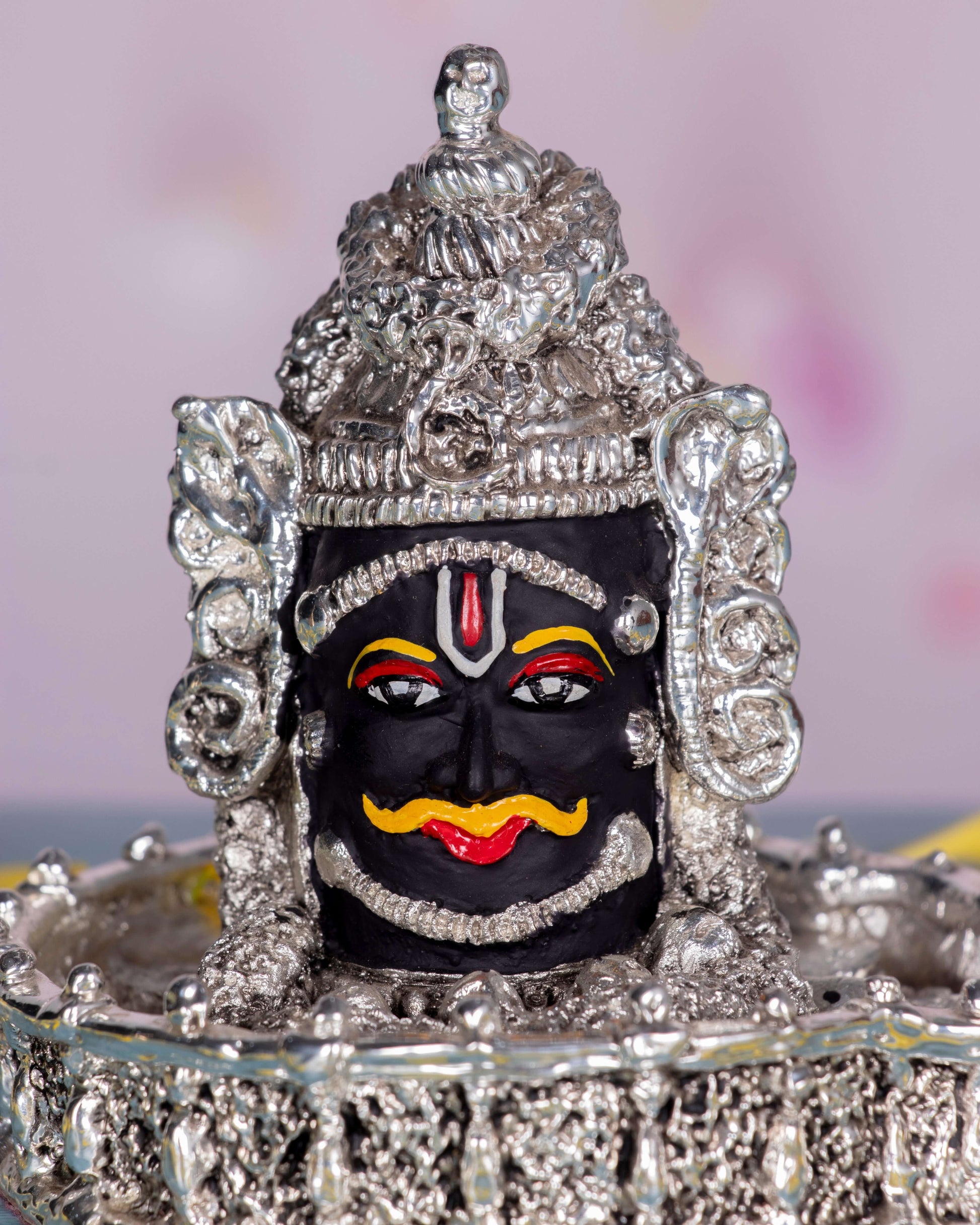 A detailed, close-up shot of a silver-plated idol of Lord Mahakal (Shiva), featuring a dark face with a prominent orange mustache, red tilak, and intricate silver ornamentation including a crown and base.