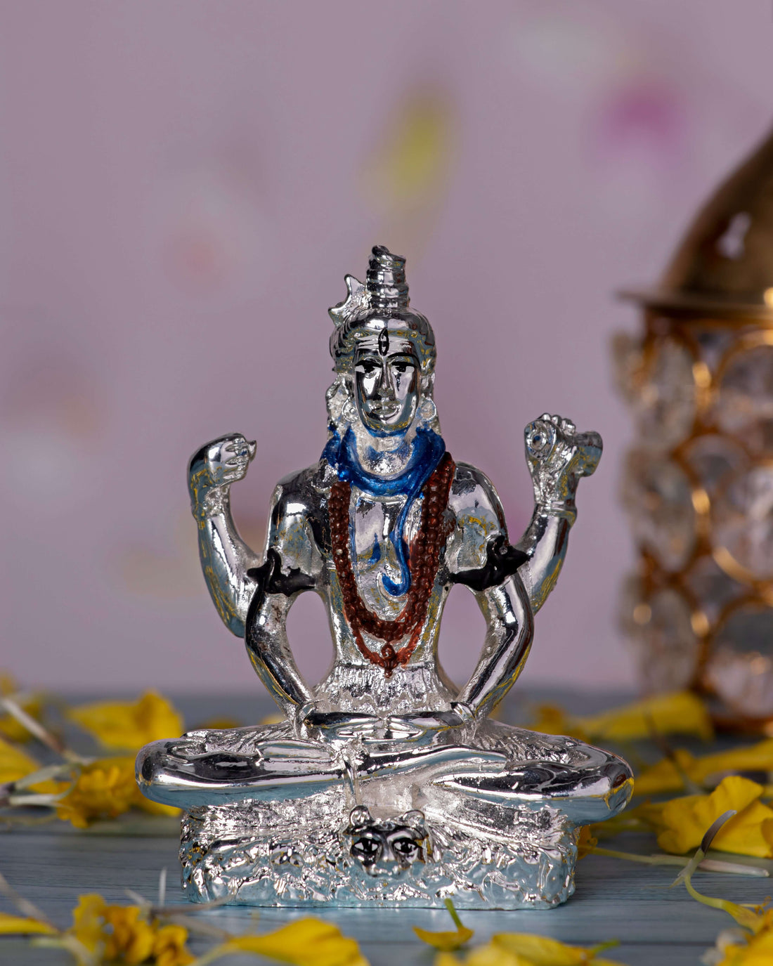 A silver-plated idol of Lord Shiva in a meditative pose, featuring a blue-painted throat and a mala around his neck, seated on a base decorated with yellow flower petals.