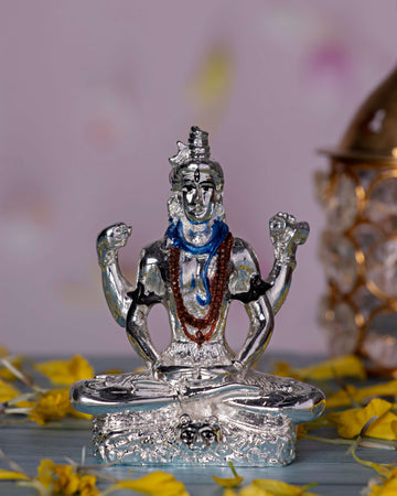A silver-plated idol of Lord Shiva in a meditative pose, featuring a blue-painted throat and a mala around his neck, seated on a base decorated with yellow flower petals.