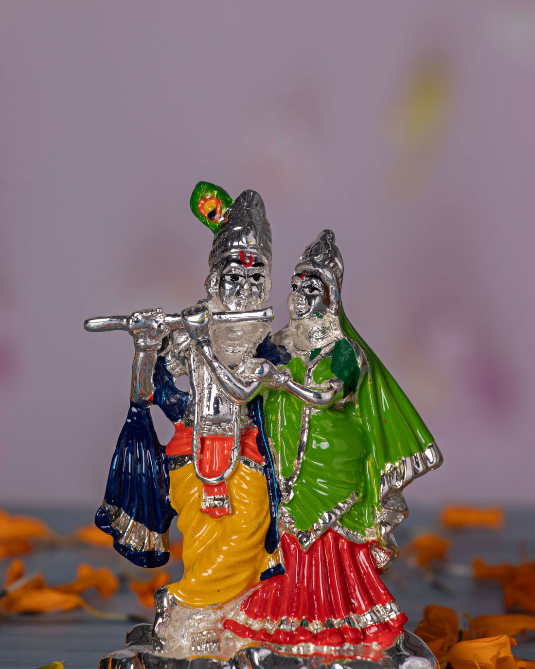 Silver-toned idol of Radha and Krishna, with Krishna playing a silver flute adorned with a green peacock feather and wearing a yellow dhoti, while Radha stands beside him in a vibrant green and red outfit. Both figures are intricately detailed and have serene expressions.
