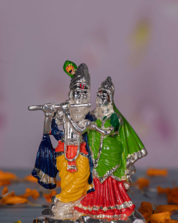Silver-toned idol of Radha and Krishna, with Krishna playing a silver flute adorned with a green peacock feather and wearing a yellow dhoti, while Radha stands beside him in a vibrant green and red outfit. Both figures are intricately detailed and have serene expressions.