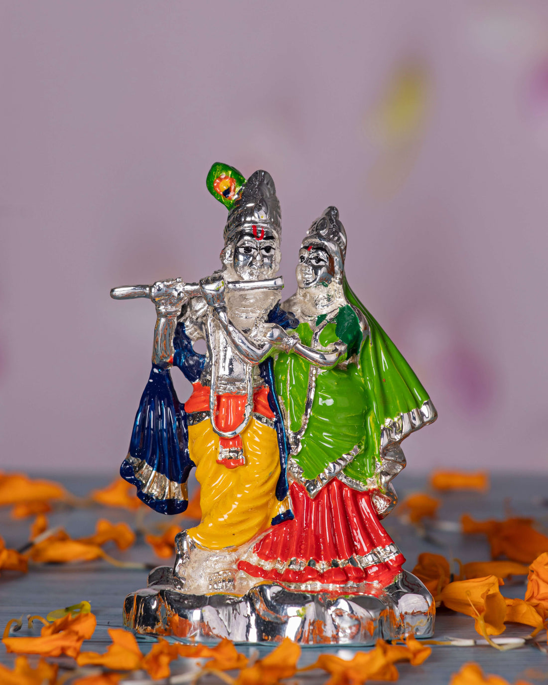 Silver-toned idol of Radha and Krishna, with Krishna playing a silver flute adorned with a green peacock feather and wearing a yellow dhoti, while Radha stands beside him in a vibrant green and red outfit. Both figures are intricately detailed and have serene expressions.