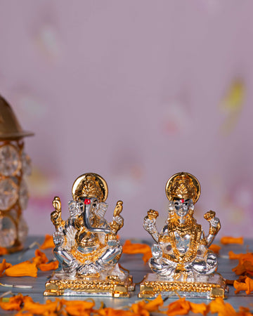 Silver and gold-plated idols of Goddess Lakshmi and Lord Ganesha seated, surrounded by orange marigold petals.