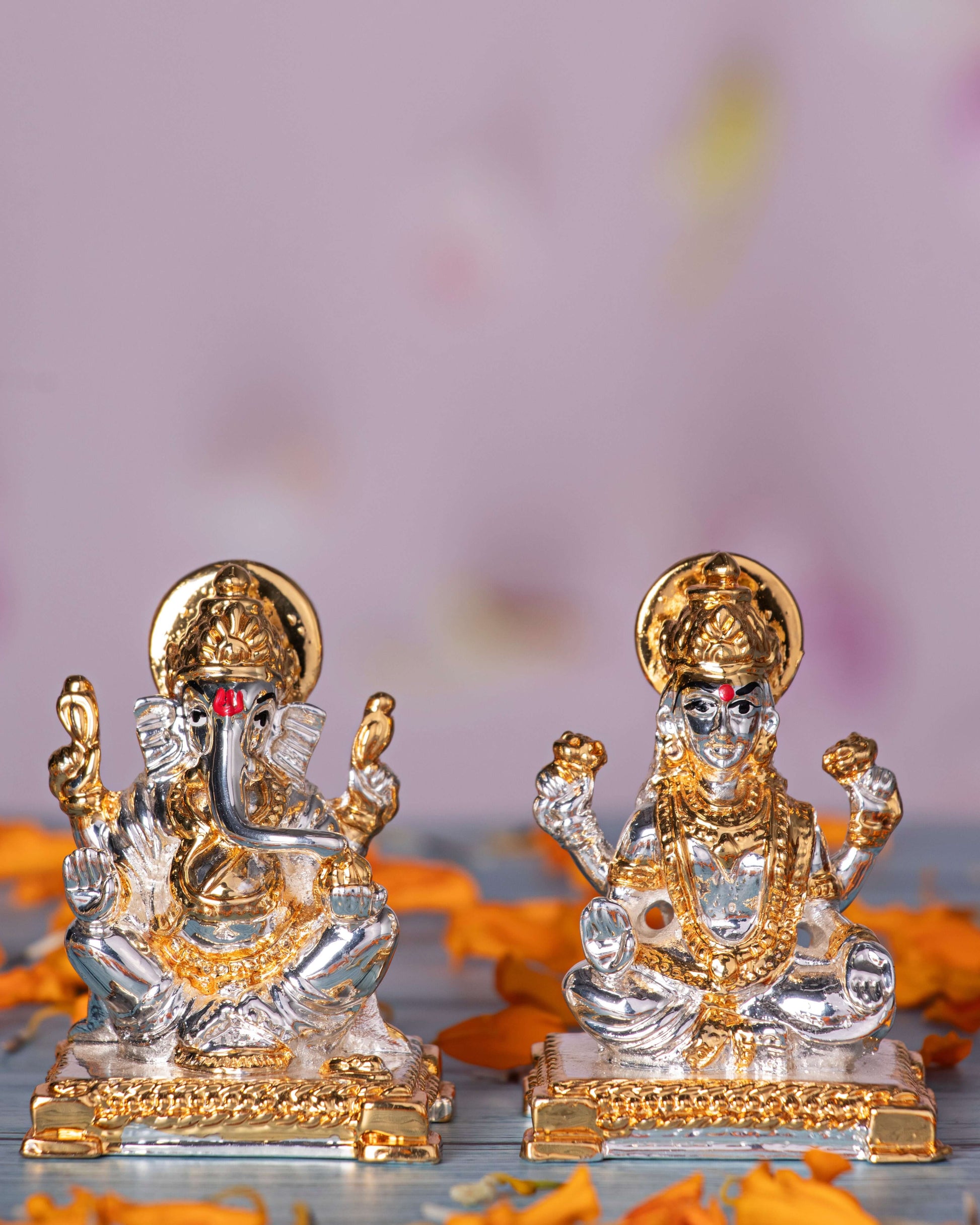Silver and gold-plated idols of Goddess Lakshmi and Lord Ganesha seated, surrounded by orange flowers.