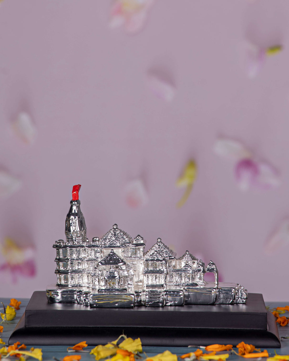 A silver idol of the Ayodhya Ram Mandir on a black base, surrounded by scattered orange and yellow flower petals on a blue wooden surface.