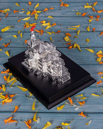 A silver idol of the Ayodhya Ram Mandir on a black base, surrounded by scattered orange and yellow flower petals on a blue wooden surface.