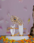 An elegant white idol depicting Lord Krishna's hands holding a golden flute, adorned with a vibrant golden peacock feather featuring a blue eye, all resting on a white base scattered with orange marigold petals.