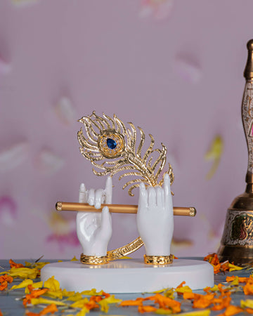 An elegant white idol depicting Lord Krishna's hands holding a golden flute, adorned with a vibrant golden peacock feather featuring a blue eye, all resting on a white base scattered with orange marigold petals.