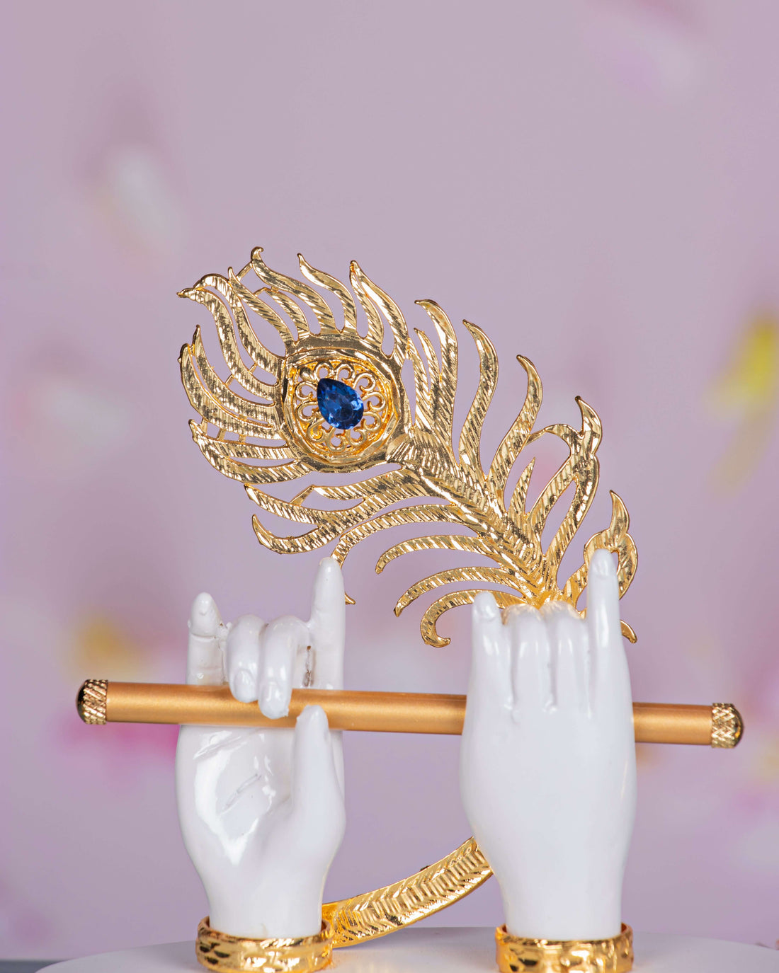 An elegant white idol depicting Lord Krishna's hands holding a golden flute, adorned with a vibrant golden peacock feather featuring a blue eye, all resting on a white base scattered with orange marigold petals.